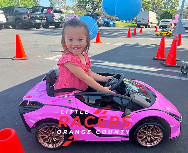 A happy toddler girl gripping the wheel of a pink branded ride-on Lamborghini on a parking-lot track