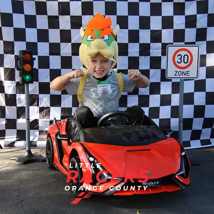 A boy in a Bowser costume hat flexing proudly in a red branded ride-on Lamborghini beside a checkered backdrop