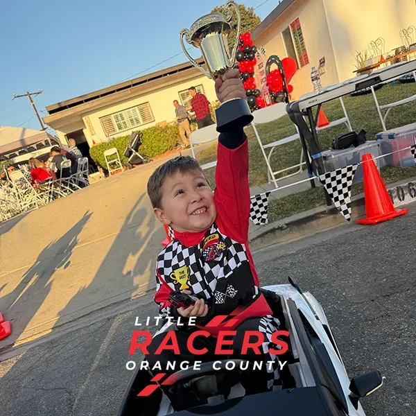 A young boy in a racing suit joyfully holding up a trophy at a Little Racers OC party at golden hour