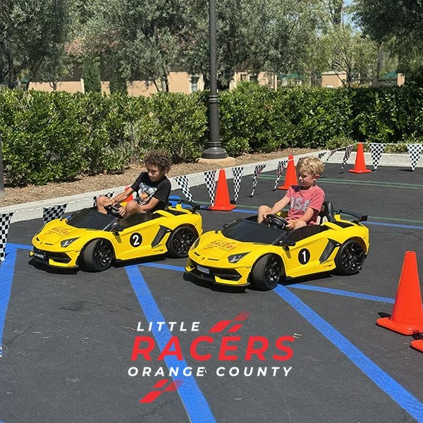 Two boys racing side by side in numbered yellow Little Racers OC ride-on Lamborghinis on a coned, checkered-flag track