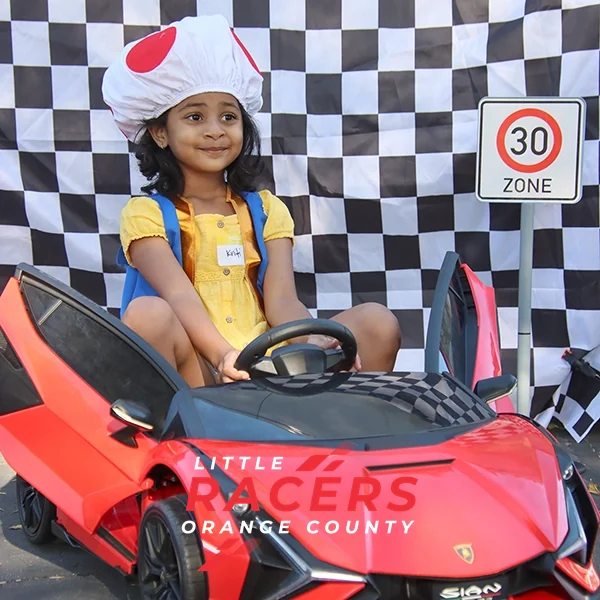 A smiling girl in a Toad costume hat sitting in a red branded ride-on Lamborghini in front of a checkered flag