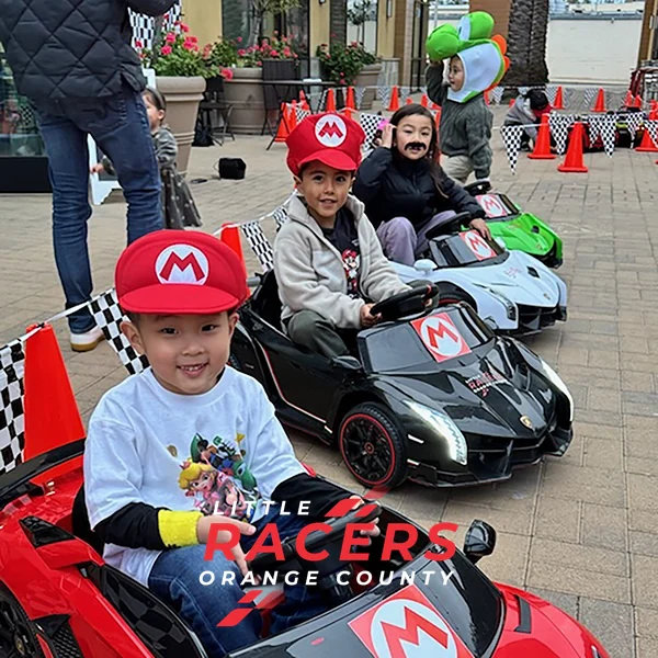 Three kids in Mario, Luigi and Peach hats racing red, black and white branded ride-on Lamborghinis at a party