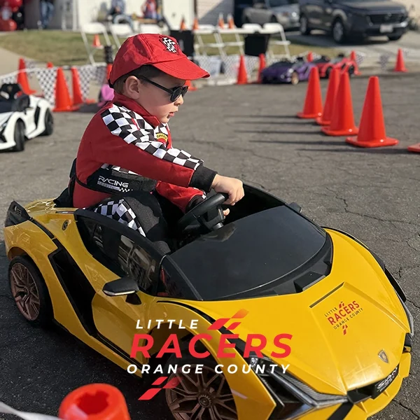 A boy in a red racing suit and cap driving a yellow Little Racers OC branded ride-on Lamborghini