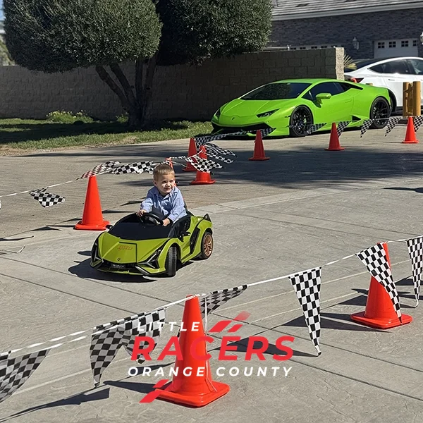 A boy driving a green ride-on Lamborghini on a checkered-flag driveway track, with a real green Lamborghini parked behind