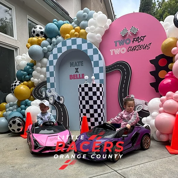 A 'Two Fast Two Curious' race-themed birthday setup with a balloon arch and pink and purple branded ride-on Lamborghinis