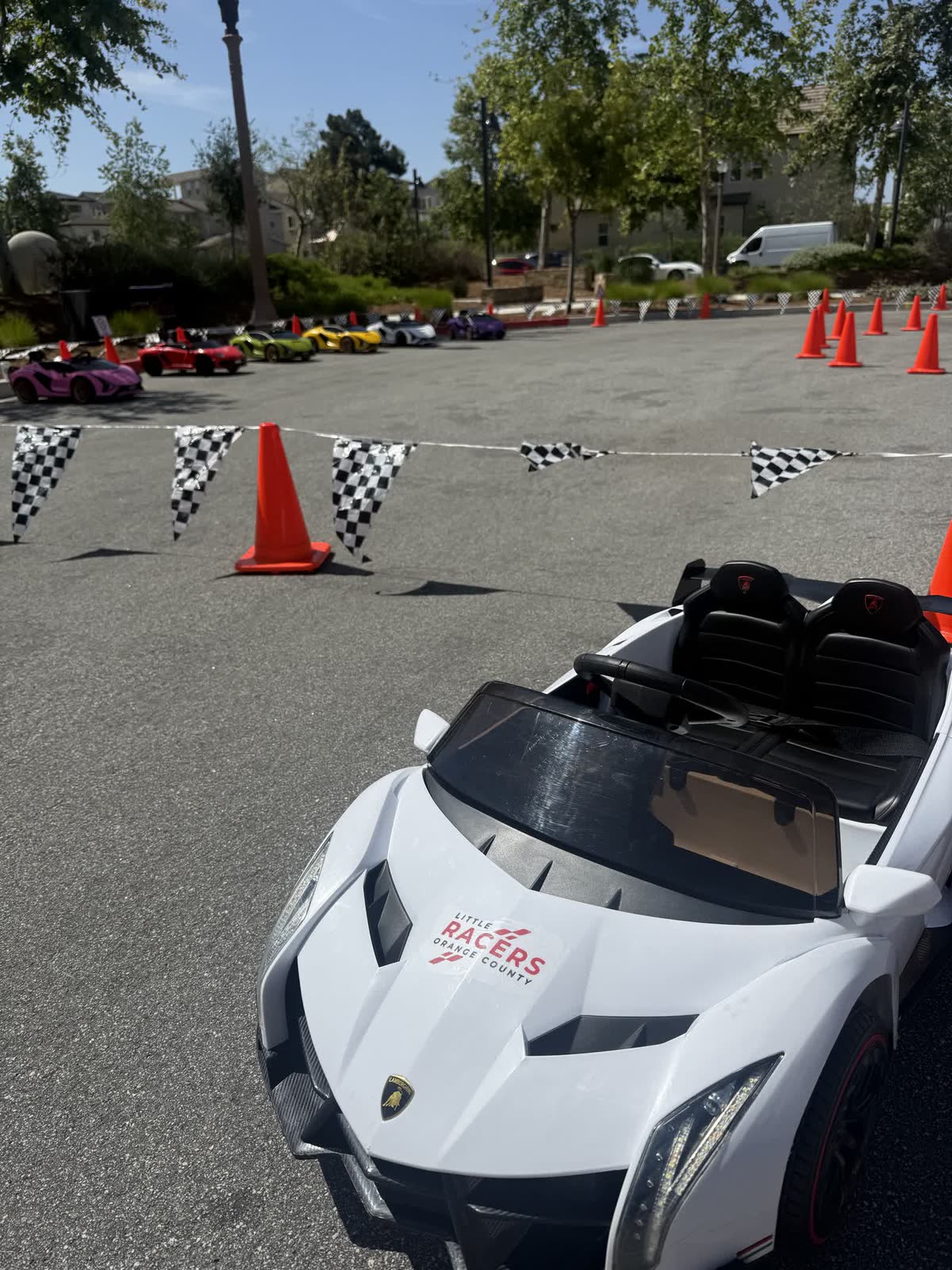 A white Little Racers OC branded kid-size Lamborghini parked in front of the full fleet of colorful ride-on cars, lined up along a checkered-flag track at an Orange County event