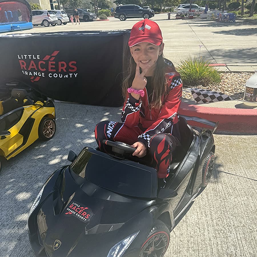 A girl in a red racing suit and cap smiling in a black Little Racers OC branded ride-on Lamborghini, beside a Little Racers Orange County banner and a yellow branded car
